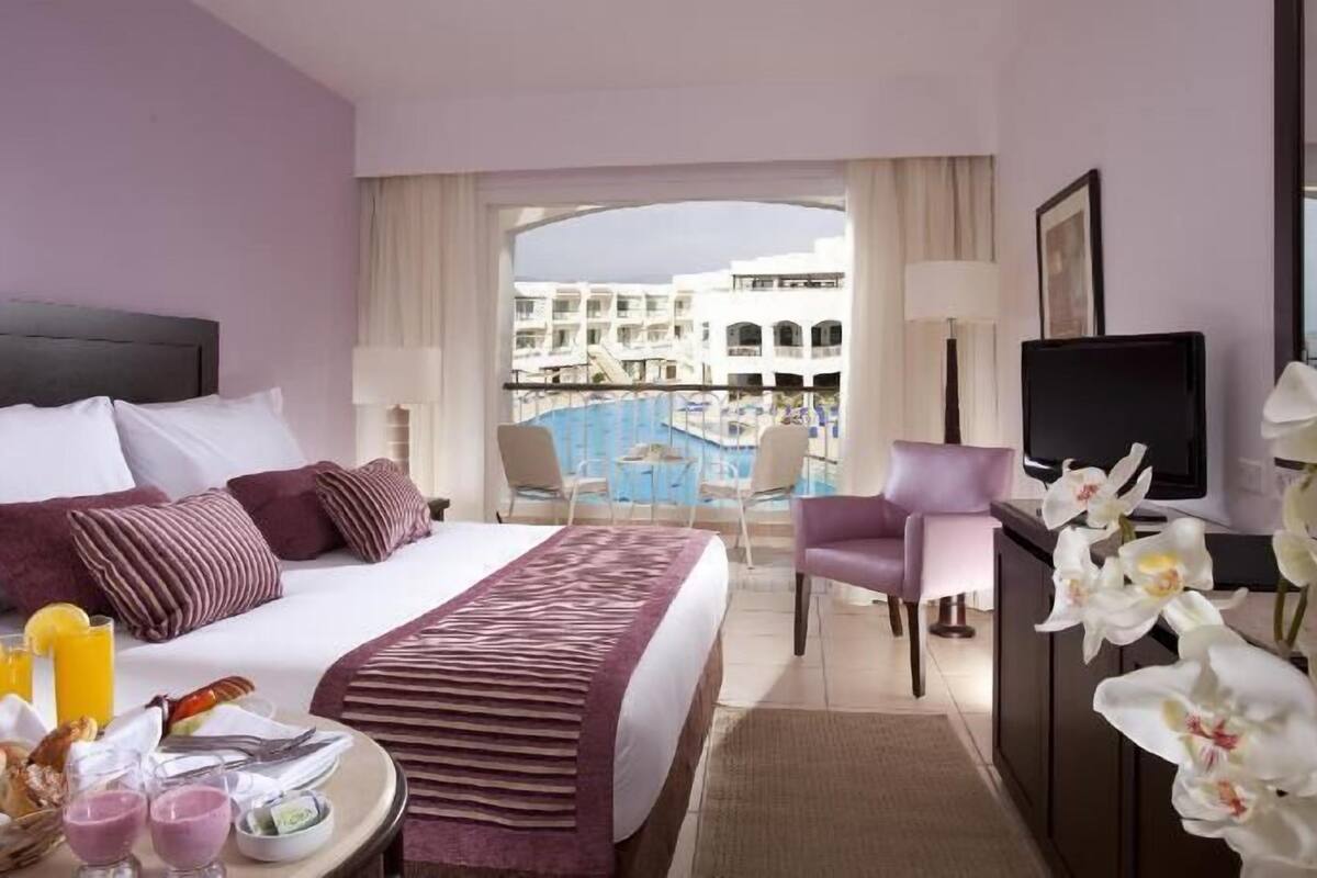 Une chambre d’hôtel équipée d’un lit, d’une télévision, d’un bureau avec des articles pour le petit-déjeuner, d’une chaise et offrant une vue sur une piscine et des bâtiments.