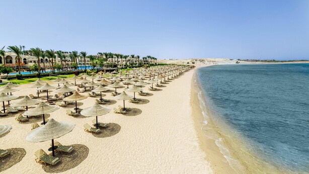 Une plage bordée de rangées de chaises longues et d’ombrelles en chaume, une piscine et, au loin, le bâtiment principal du complexe hôtelier.