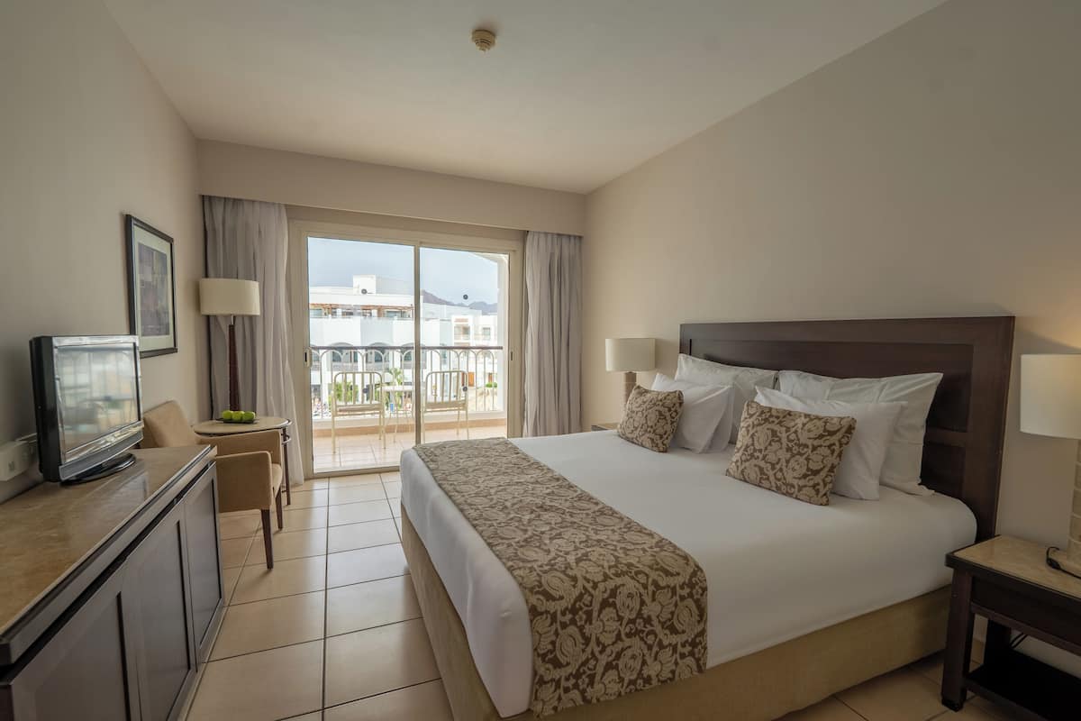 Une chambre d’hôtel avec un grand lit, une télévision, un bureau et un balcon offrant une vue sur les bâtiments et les montagnes.