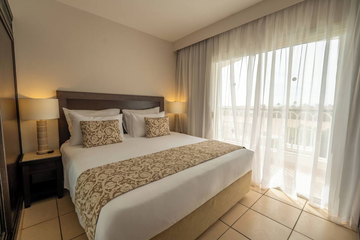 Une chambre d’hôtel avec un grand lit, deux lampes de chevet, une couvre-lit à motifs et des rideaux légers donnant sur un balcon.