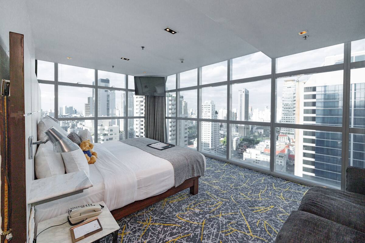 A high-rise hotel room with a large bed, a bedside lamp, a bedside table with a phone and notepad, a teddy bear, and a city view through floor-to-ceiling windows.