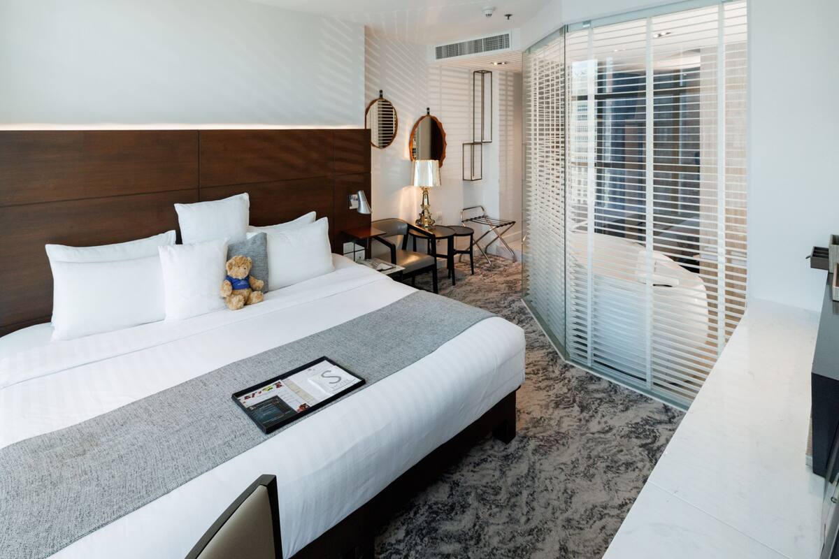 A hotel room with a large bed, a desk, a chair, a small table, and a window with blinds.