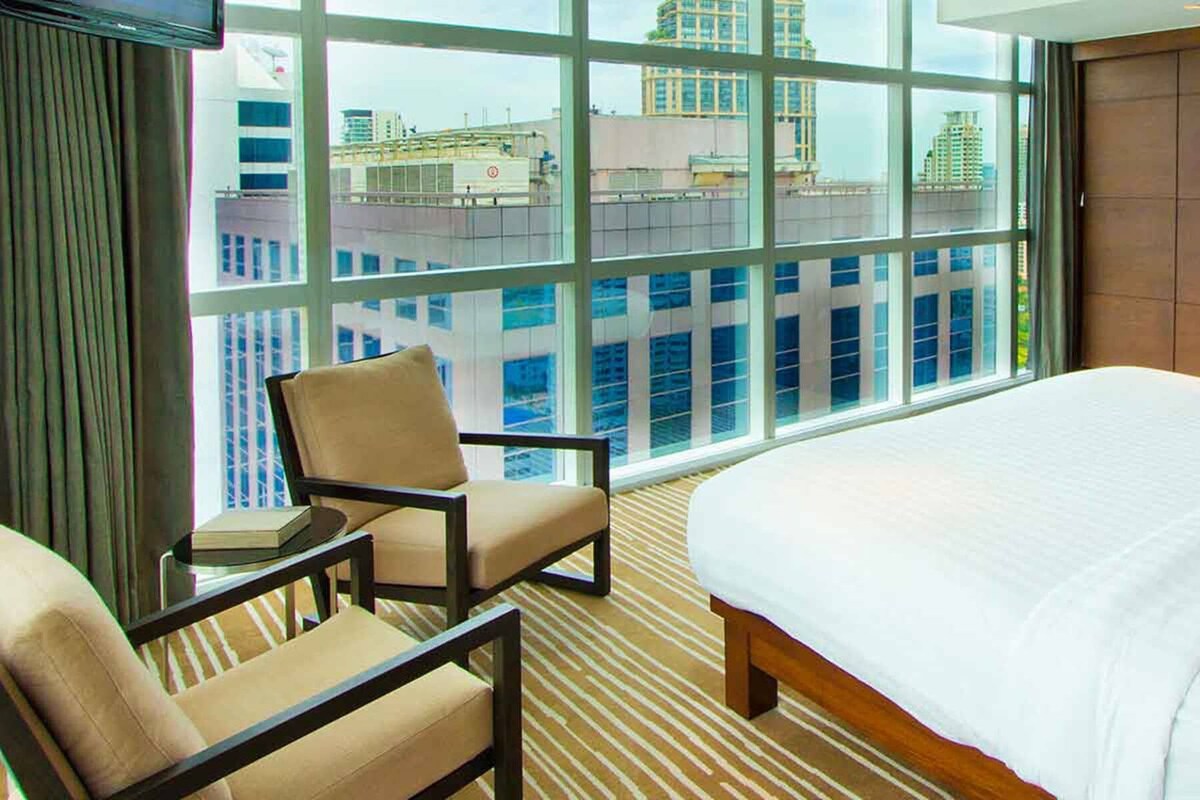 A hotel room with a large bed, a chair, a small table, a microwave, and a view of the city through large windows.