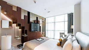 Una habitación de hotel moderna con una cama grande, un televisor de pantalla plana montado en la pared, una mesita de noche con lámpara, un ventanal con cortinas y un baño con lavabo de mármol.