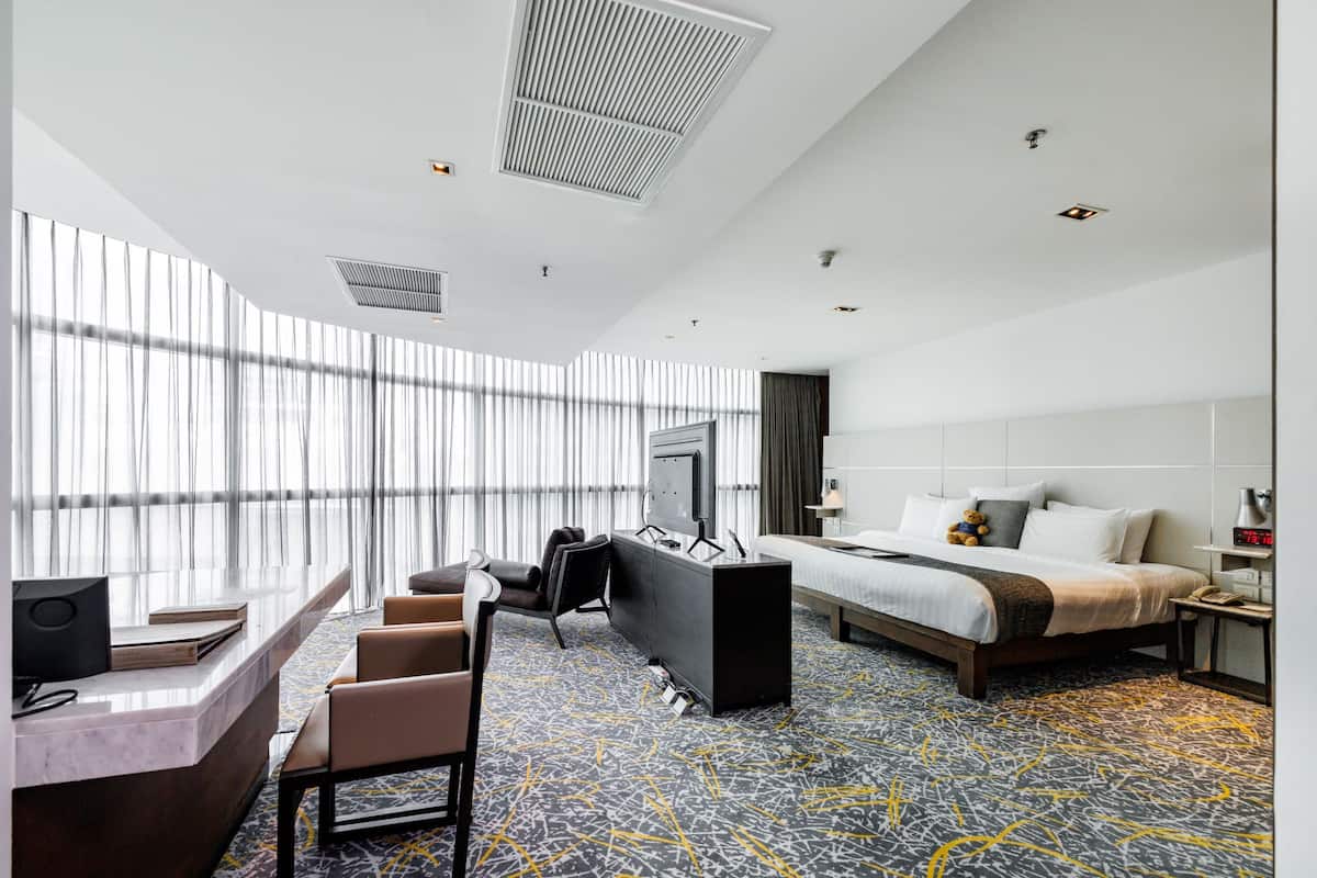 A hotel room with a large bed, a desk with a chair, a TV, and a large window with curtains.