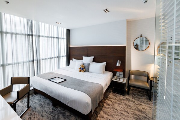 Habitación de hotel con una cama grande, una mesita de noche con lámpara, una silla y vistas a la ciudad a través de amplios ventanales.