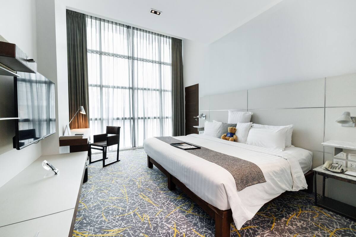 A hotel room with a large bed, a desk, a chair, a TV, and a window with curtains.