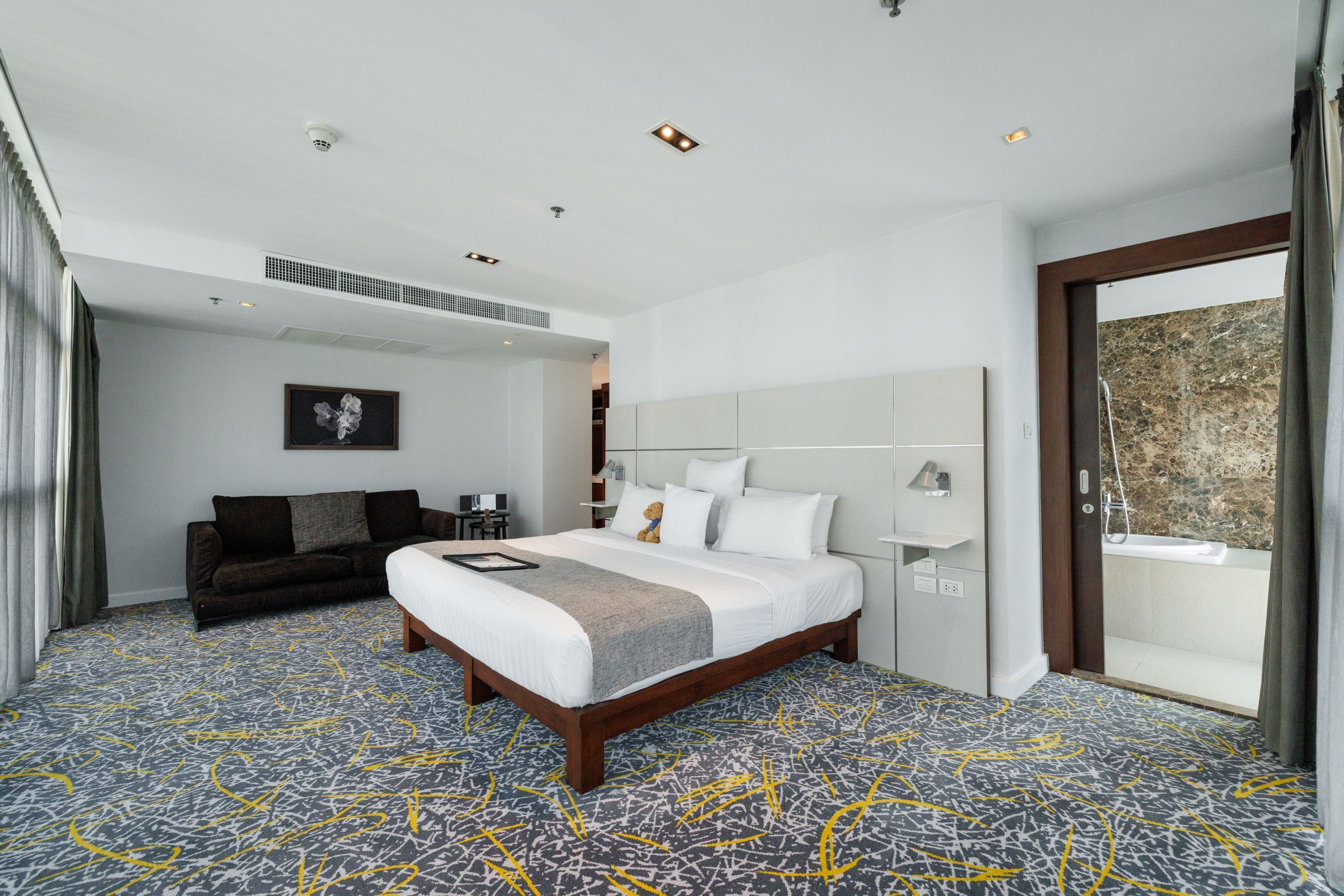 A modern hotel room with a large bed, a sofa, a bathroom with a bathtub, and a view of the outside.