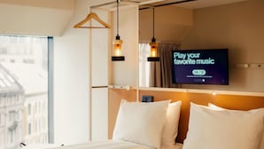 A hotel room with a bed, a TV mounted on the wall, and a window with a view of buildings. - Scandic Go, Grensen 20 (Oslo)