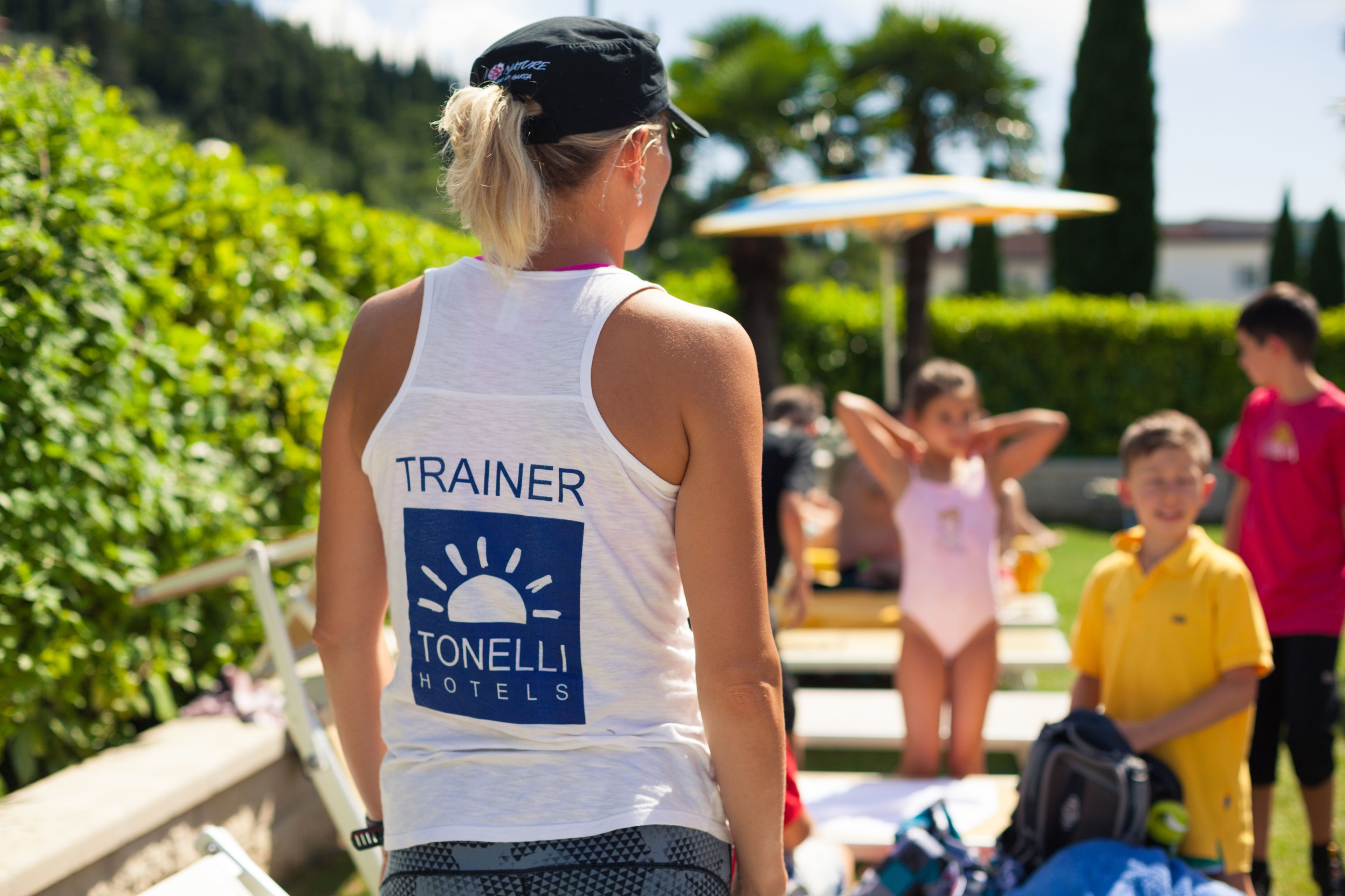 A person wearing a "TRAINER TONELLI HOTELS" shirt, standing outdoors with greenery and a clear sky in the background.
