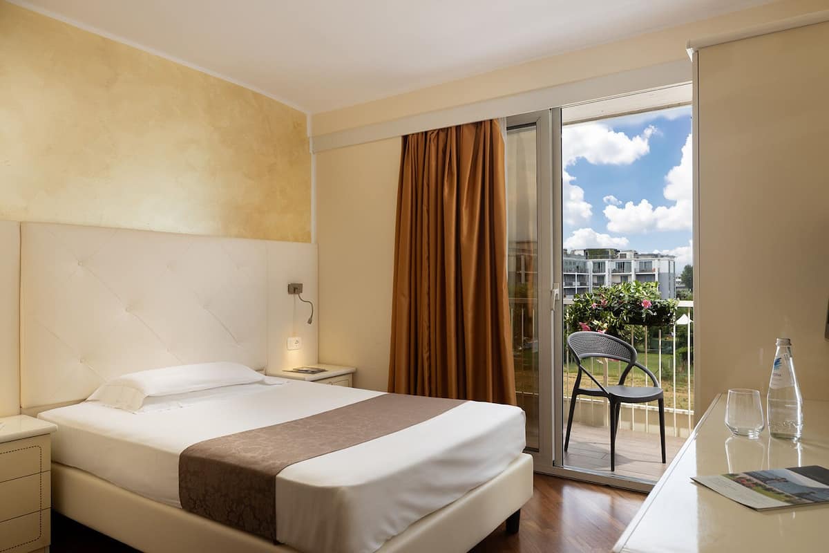 A hotel room with a bed, a nightstand, a desk with a chair, a window with curtains, and a view of buildings and greenery outside.