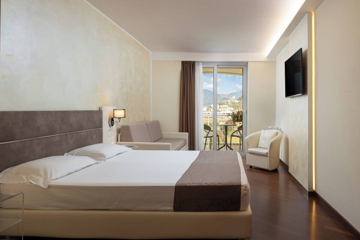 A hotel room with a large bed, a sofa, a chair, a TV, and a balcony with a view.