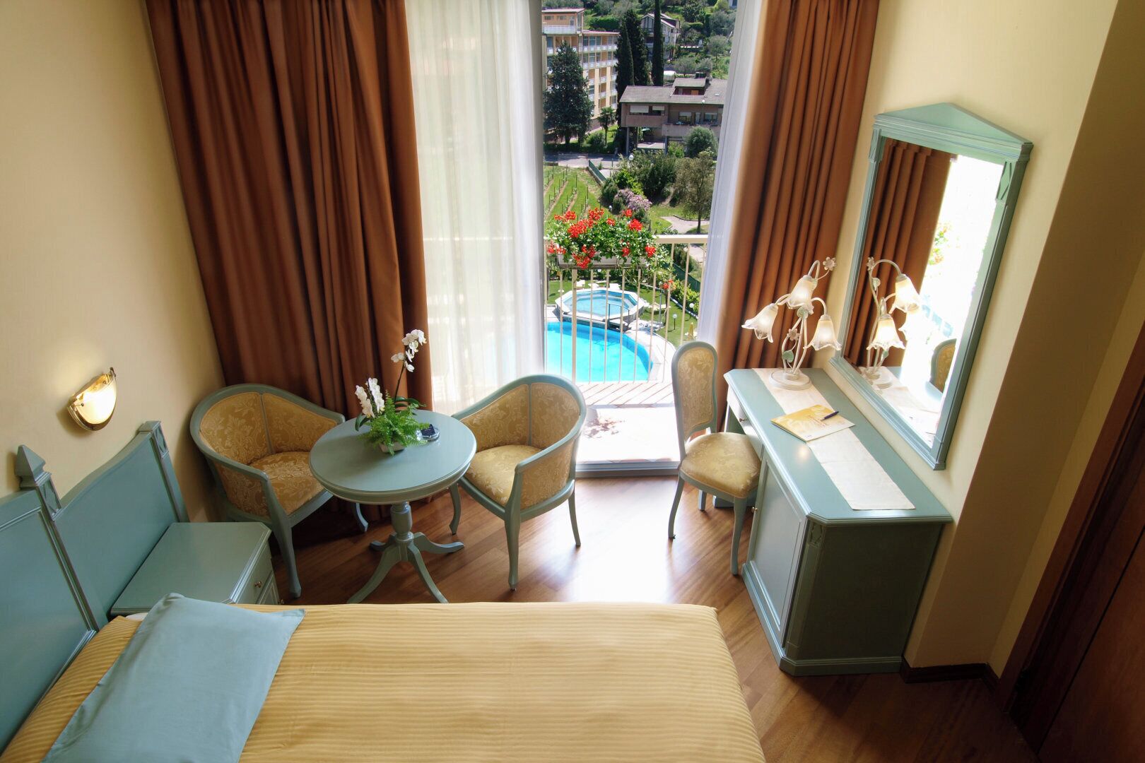 Ein Hotelzimmer mit einem Bett, zwei Sesseln, einem kleinen runden Tisch mit einer Pflanze, einem Schreibtisch mit einer Lampe und Blick auf einen Pool und Grünflächen.