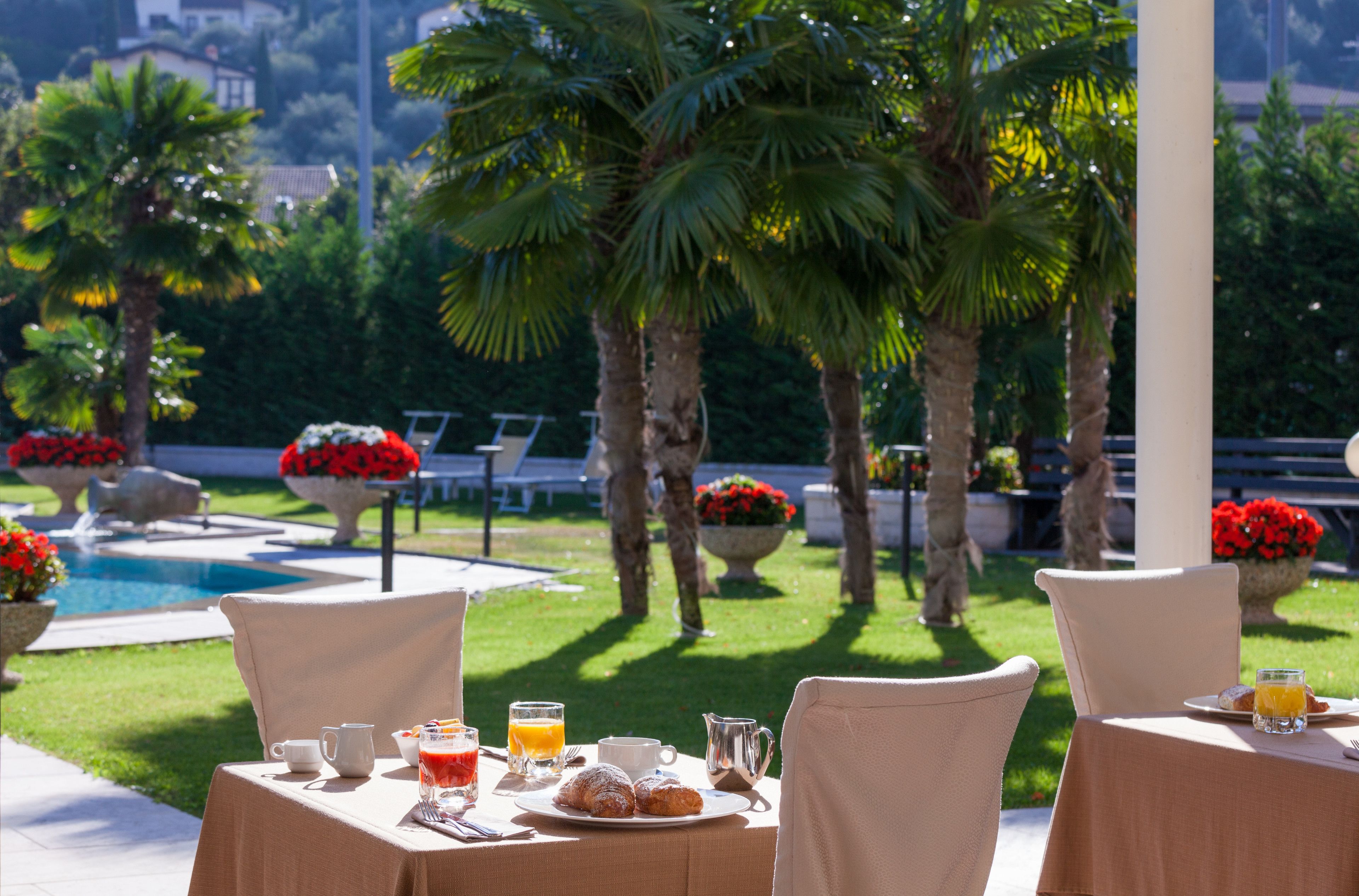 An outdoor dining area with tables set for breakfast, a pool, and palm trees in the background.