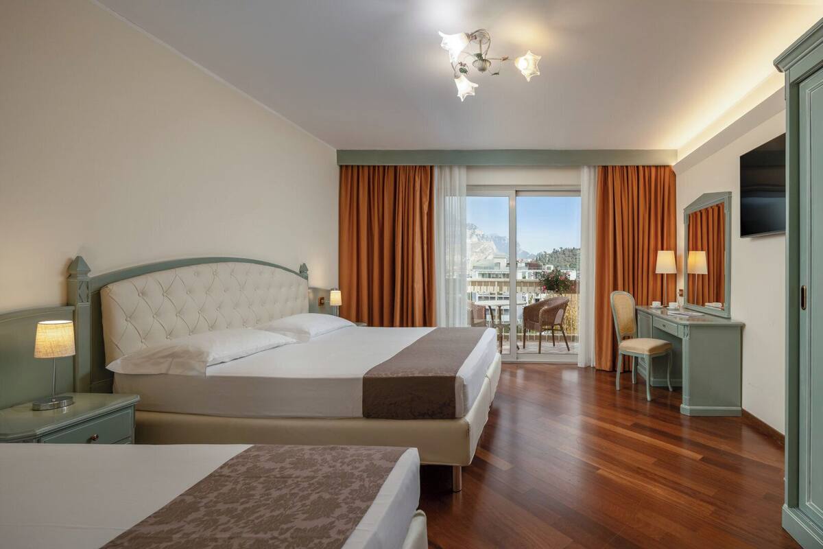 A hotel room with two beds, a desk, a chair, a television, and a balcony with curtains.