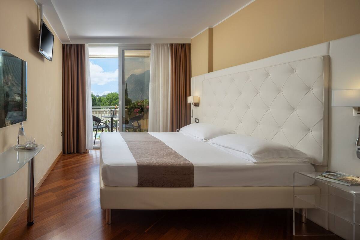 A hotel room with a large bed, a TV mounted on the wall, a glass table with a bottle and glasses, and a view of mountains through the window.