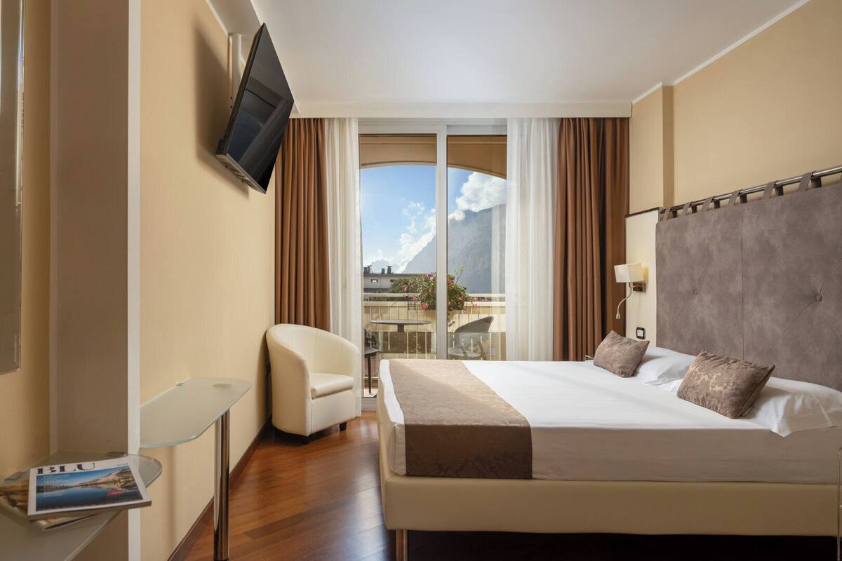 A hotel room with a large bed, a TV mounted on the wall, a chair, and a balcony with a view of the mountains.
