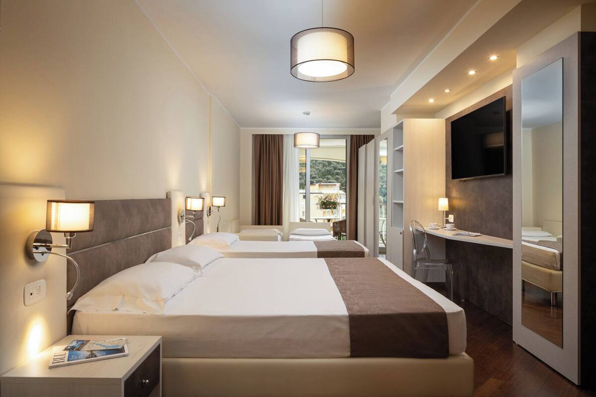 A hotel room with two beds, a desk, a TV, and a balcony with a view.