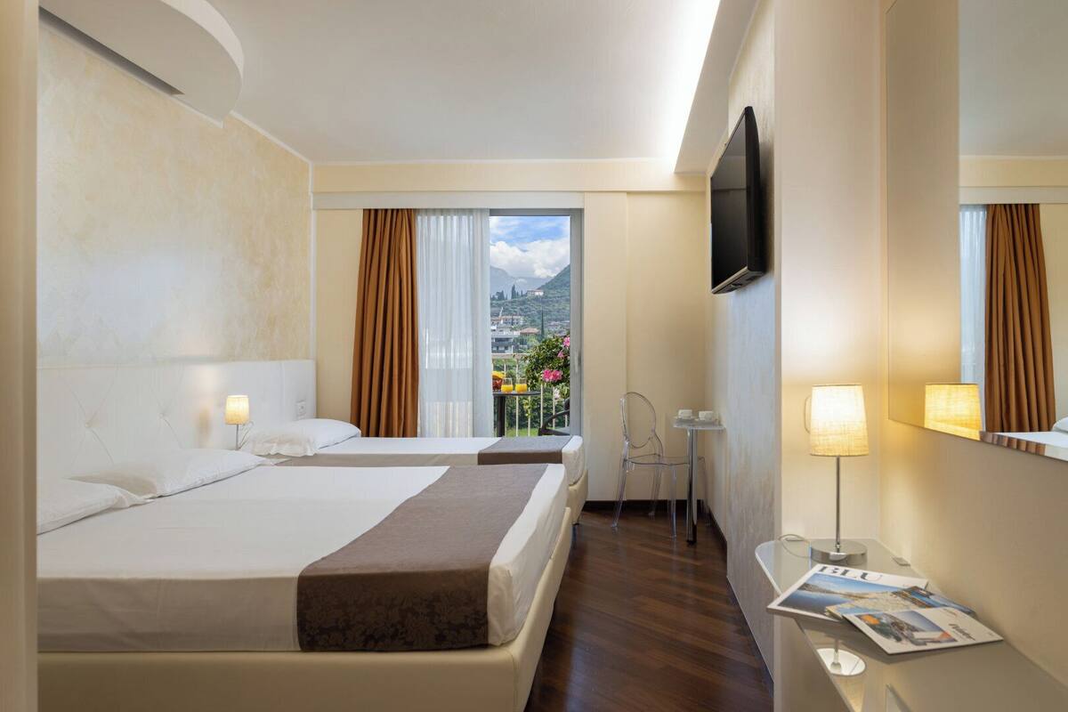 A hotel room with two beds, a balcony with a city view, a TV mounted on the wall, and a small table with a lamp.