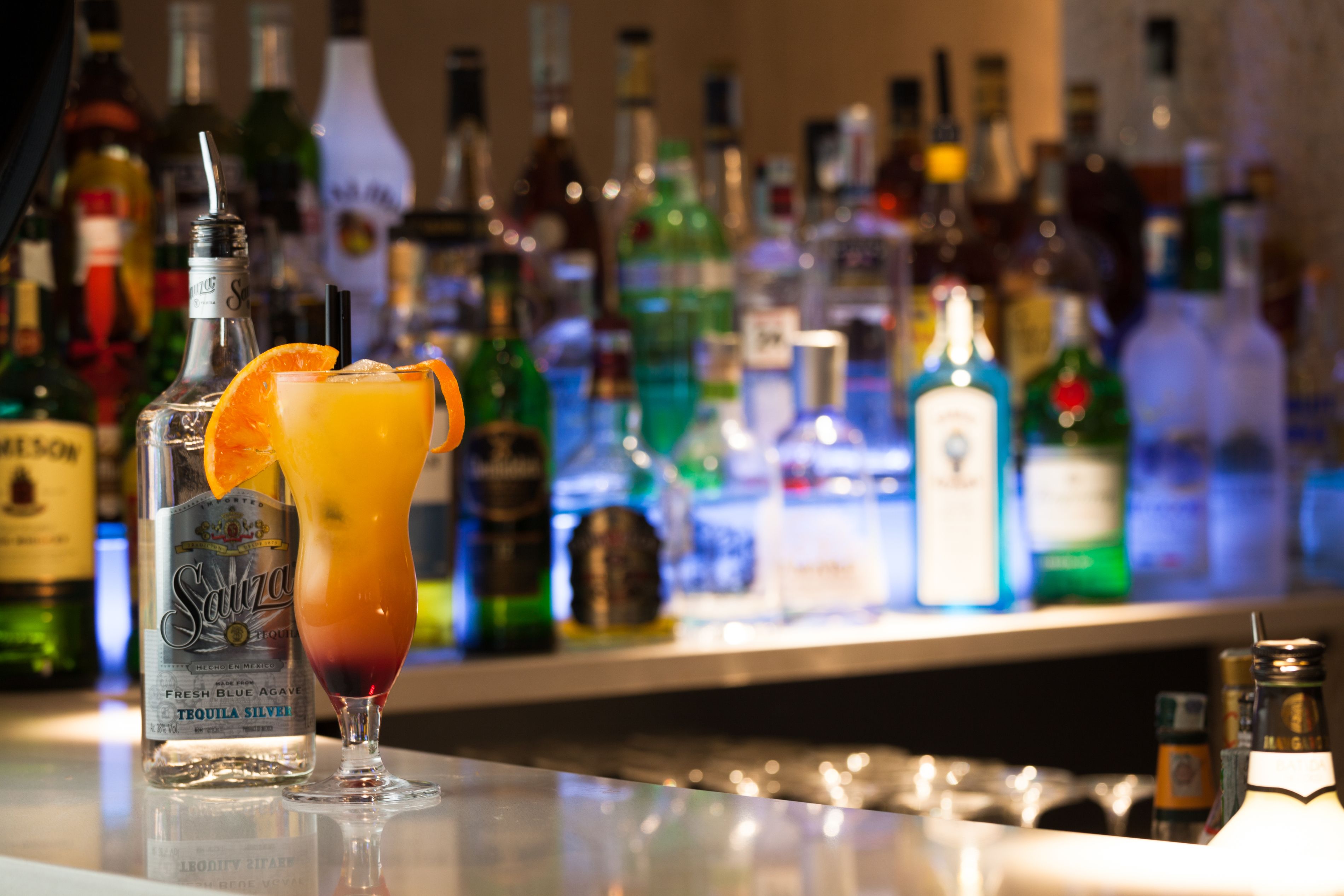 Un bar con una variedad de botellas de licor al fondo y un cóctel con un adorno de naranja sobre la barra.