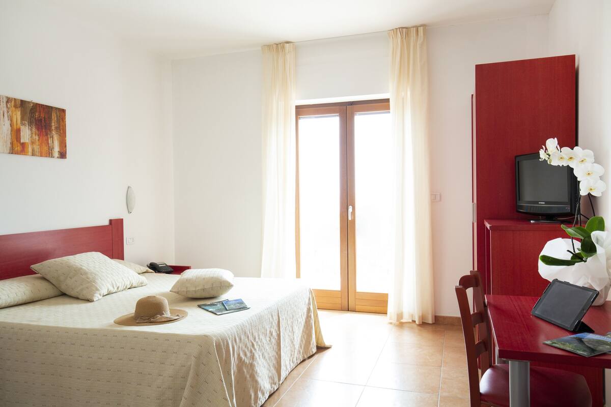 Une chambre d’hôtel avec un lit, un bureau avec un ordinateur portable, une télévision et une fenêtre avec des rideaux.