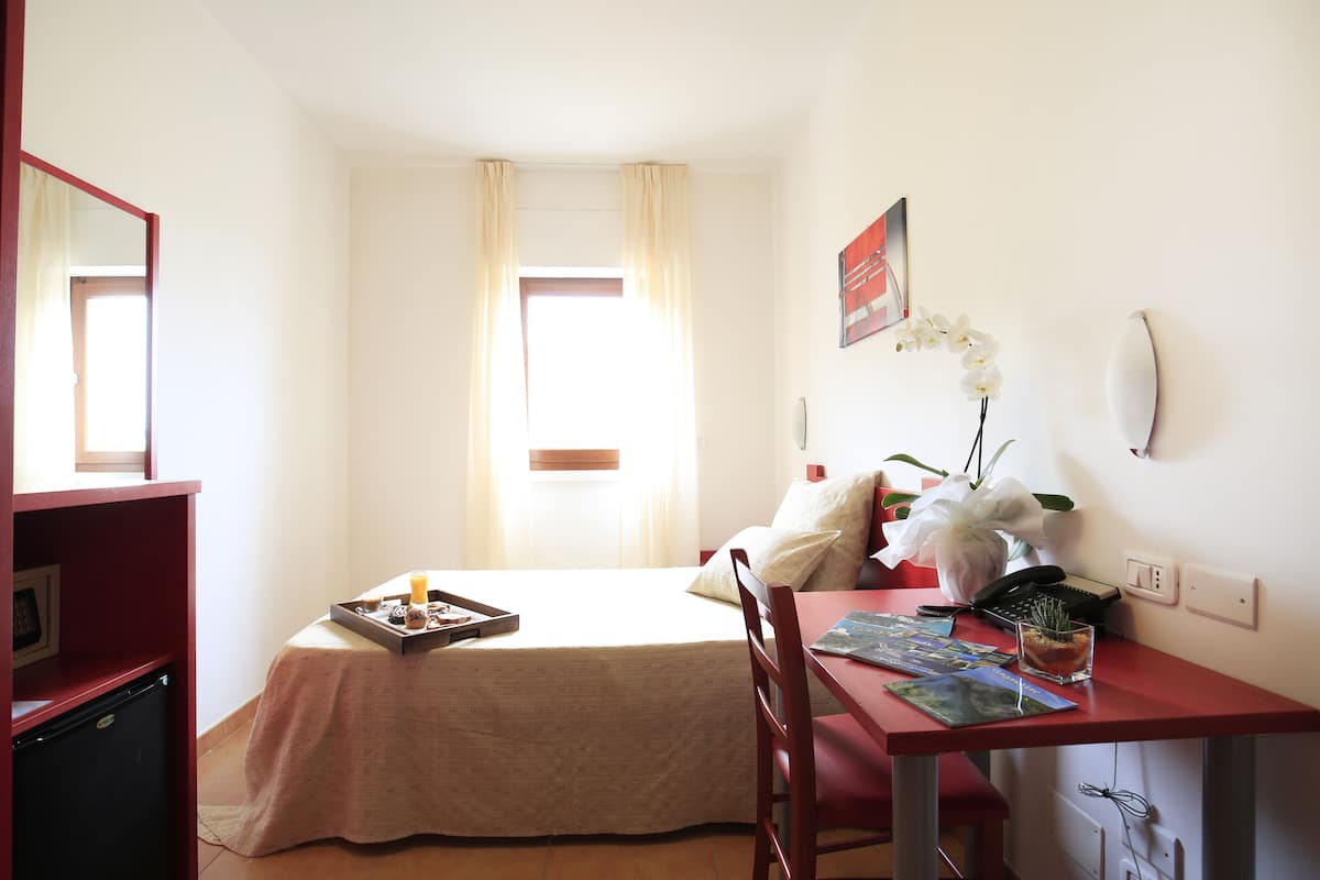 Une chambre d’hôtel comprenant un lit, un bureau avec une chaise, un petit réfrigérateur, une fenêtre avec des rideaux et un téléphone fixé au mur.