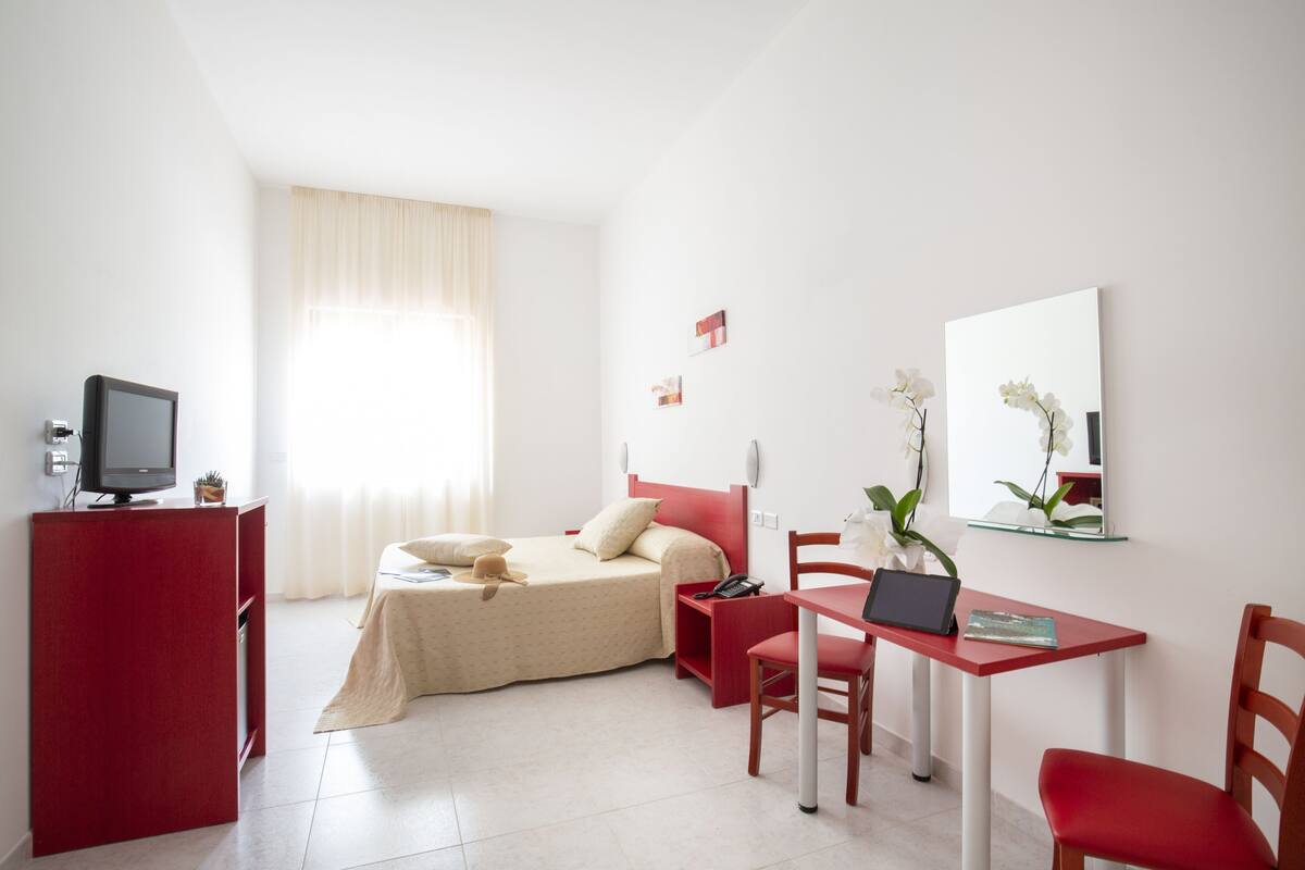 Une chambre d’hôtel avec un lit, un bureau avec un ordinateur portable, une télévision et un fauteuil rouge.