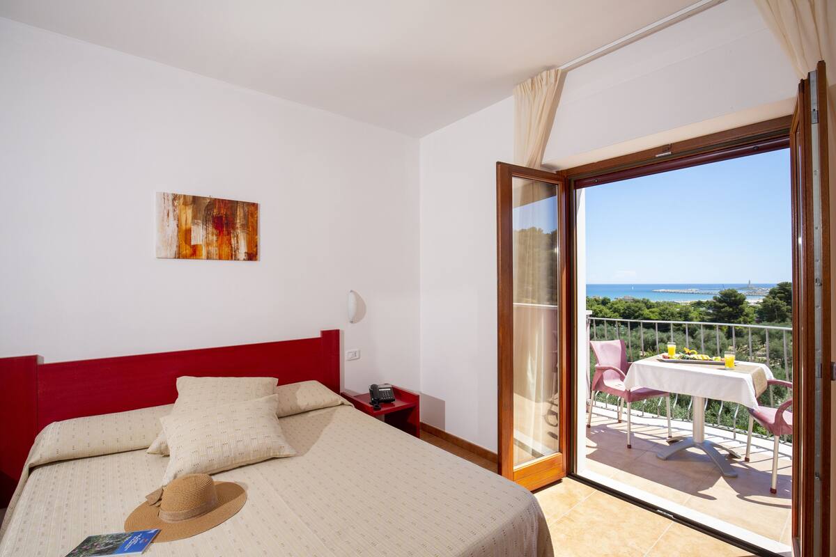 Une chambre d’hôtel avec un lit, une tête de lit rouge, un tableau au mur, une table de chevet, un téléphone et un balcon avec une table et des chaises.