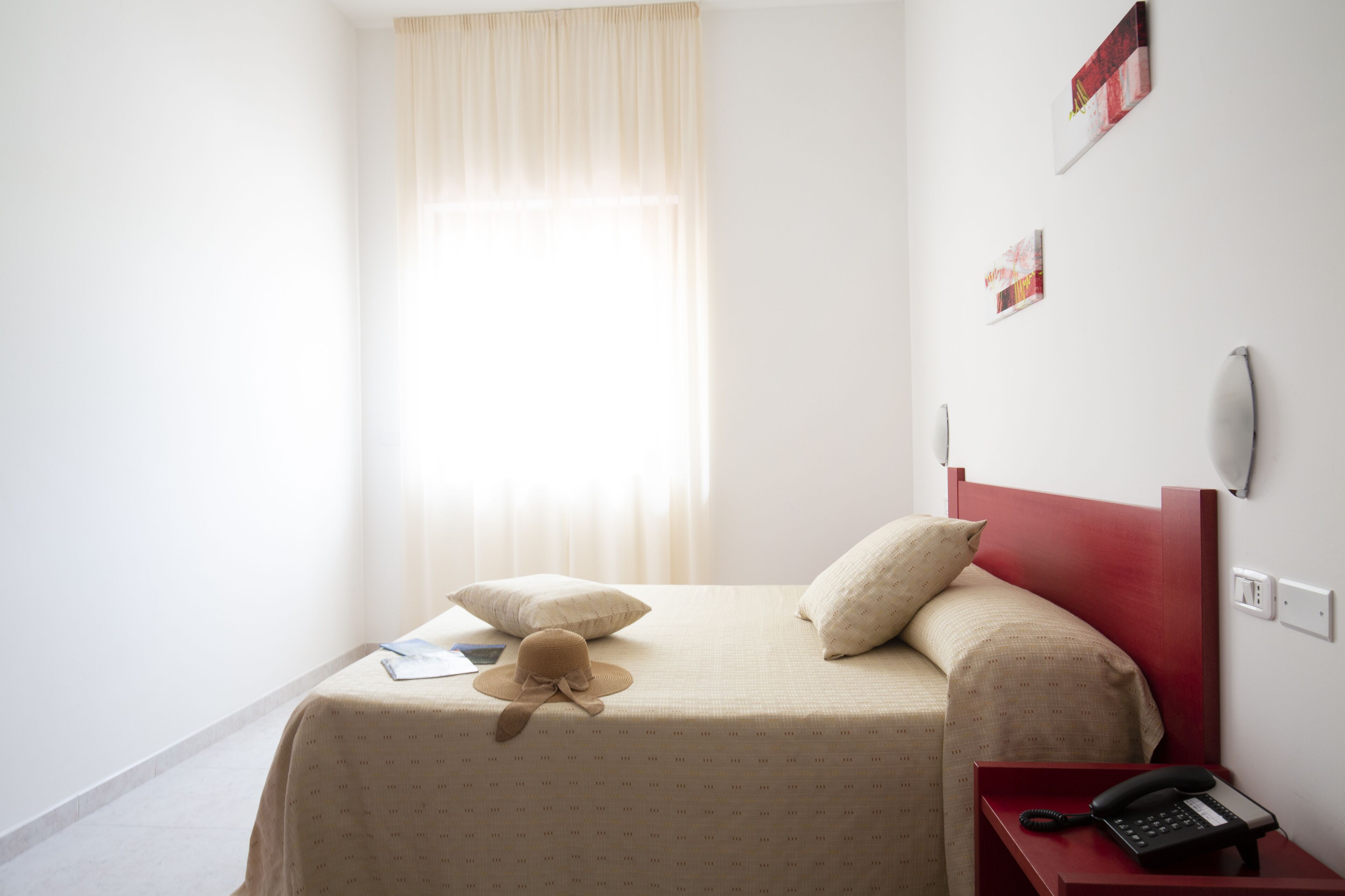 A hotel room with a bed, a nightstand with a phone, a window with curtains, and a wall with a light switch.