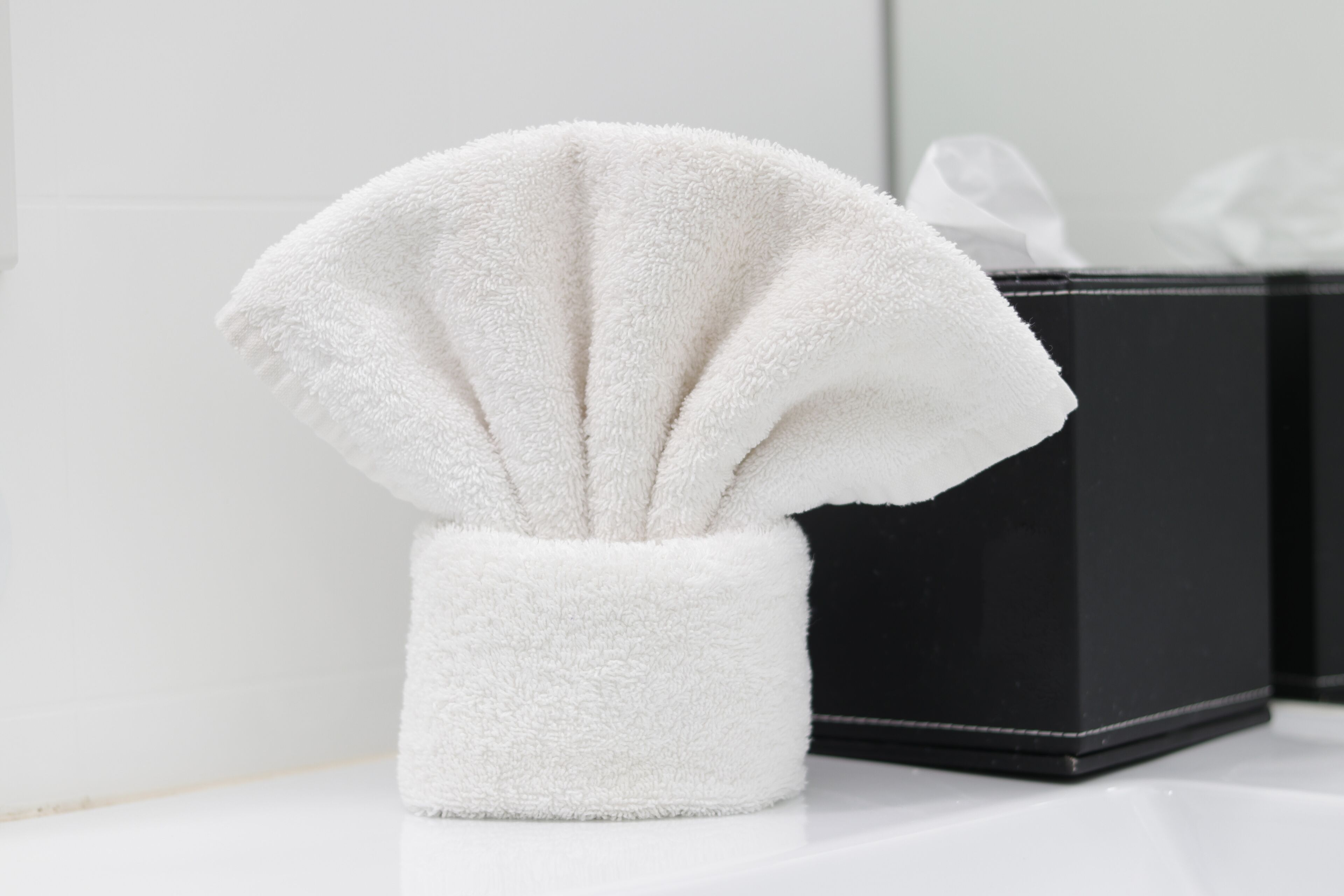 Een witte handdoek in een witte houder op een wit oppervlak, met een zwarte doos op de achtergrond.