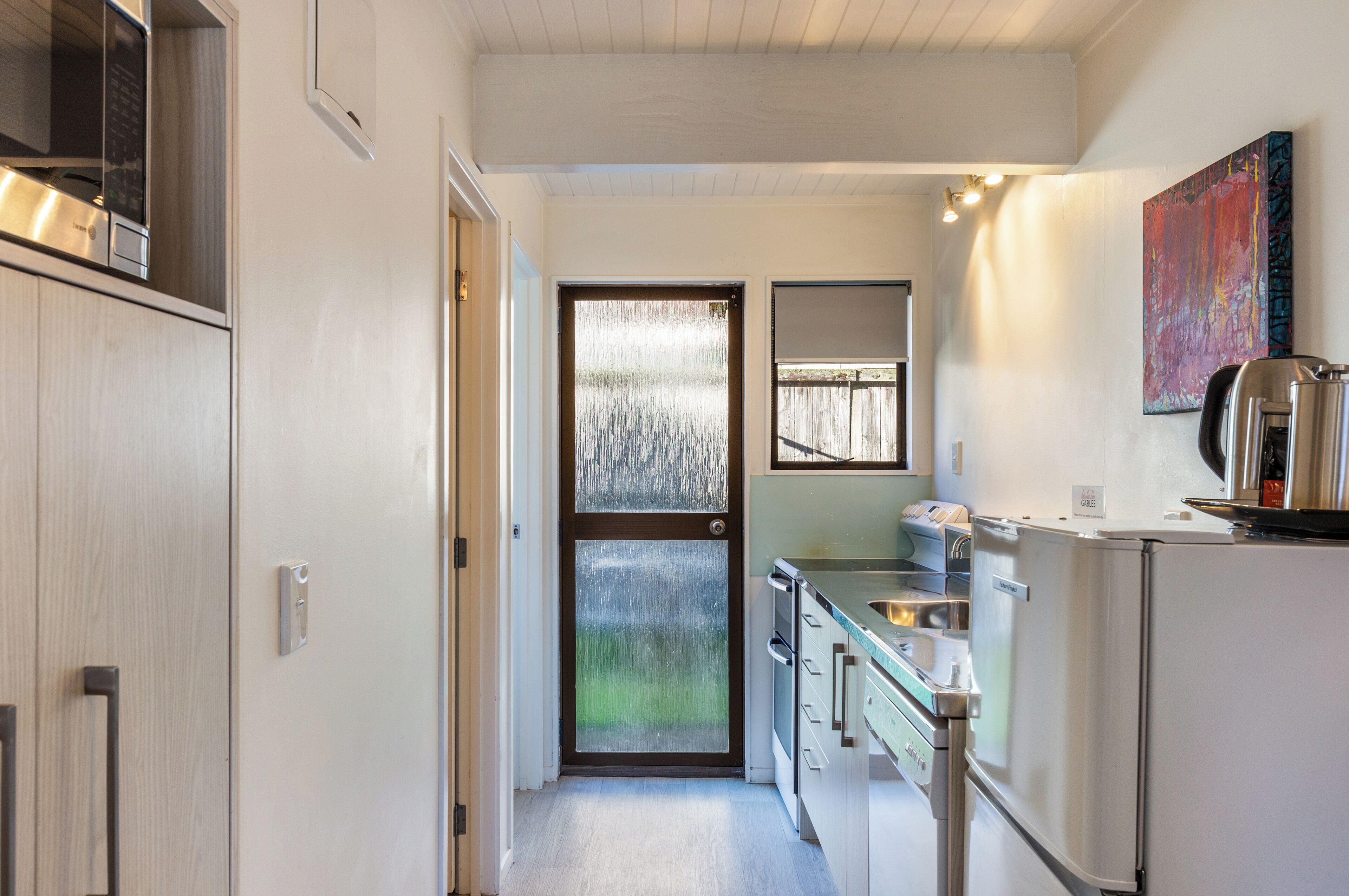 A modern kitchen with stainless steel appliances, a sliding glass door leading to a balcony, and a colorful abstract painting on the wall.