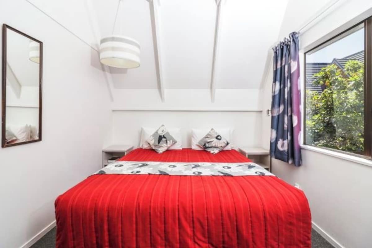 A bedroom with a red bedspread, white walls, a large window with curtains, and a ceiling light fixture.