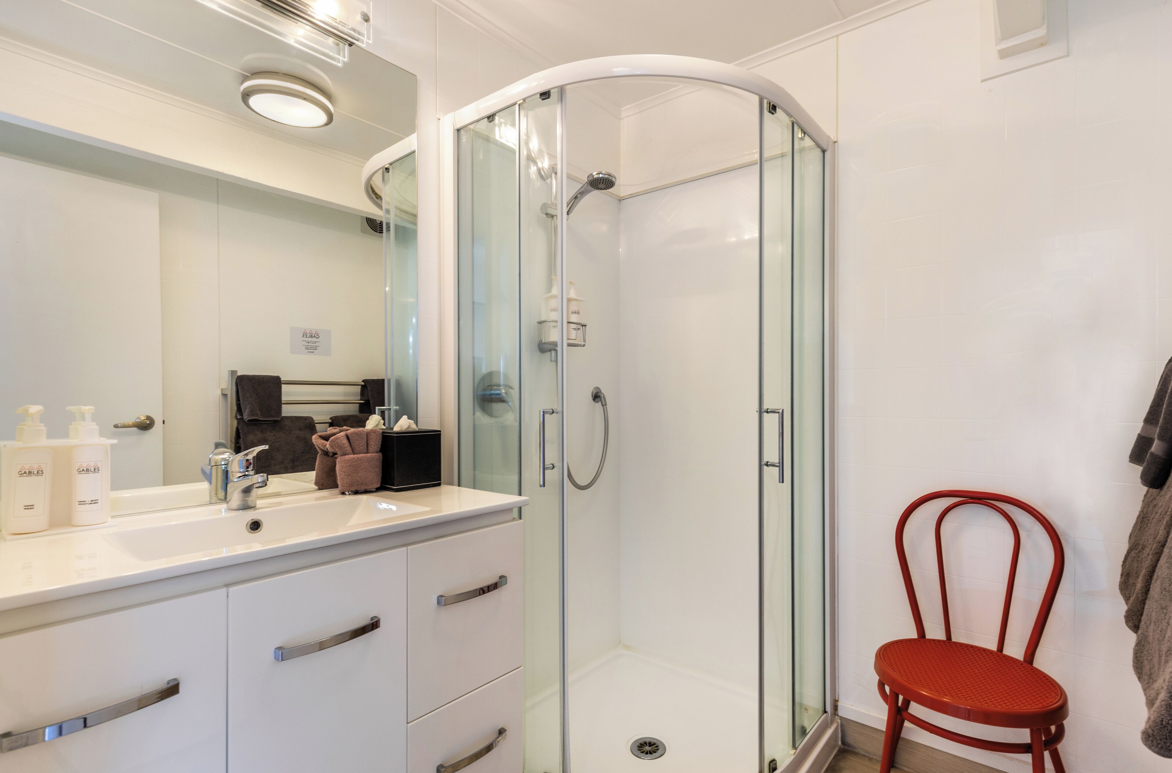 A modern bathroom with a glass-enclosed shower, a white vanity with a mirror, and a red chair.