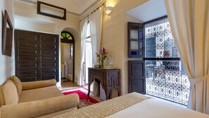 Une chambre à coucher avec un grand lit, un canapé, une petite table sur laquelle est posé un vase, et une fenêtre ornée de barreaux en fer décoratifs.