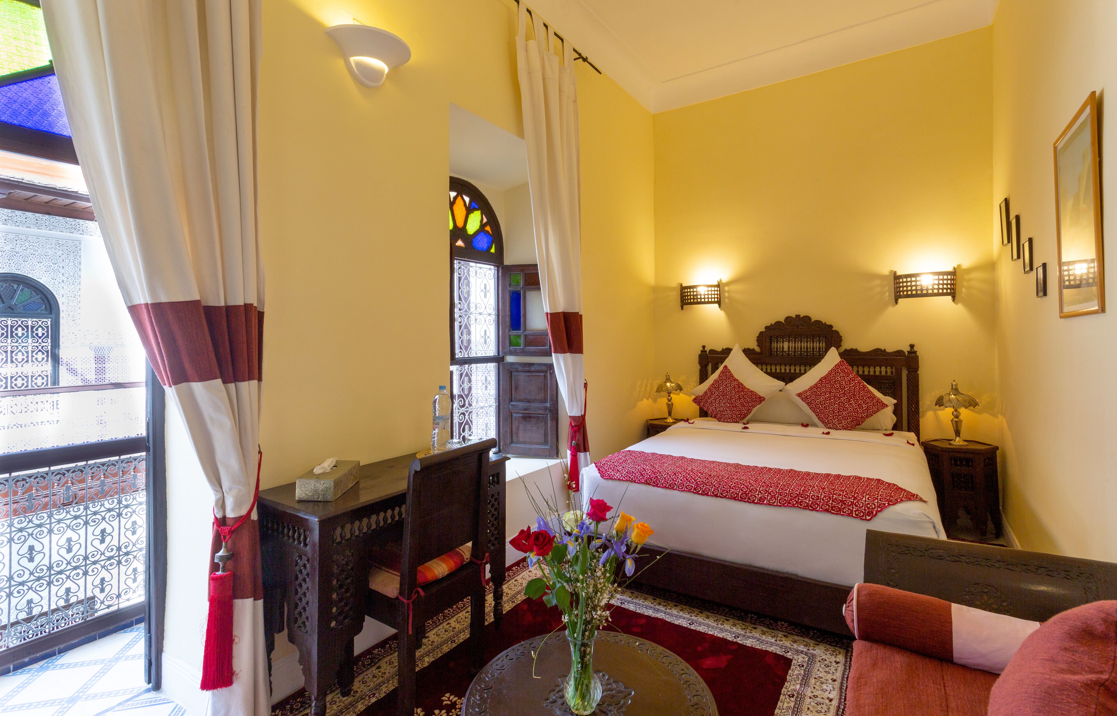 A hotel room with a bed, bedside tables, a chair, a small table with a vase of flowers, and a balcony with a railing.