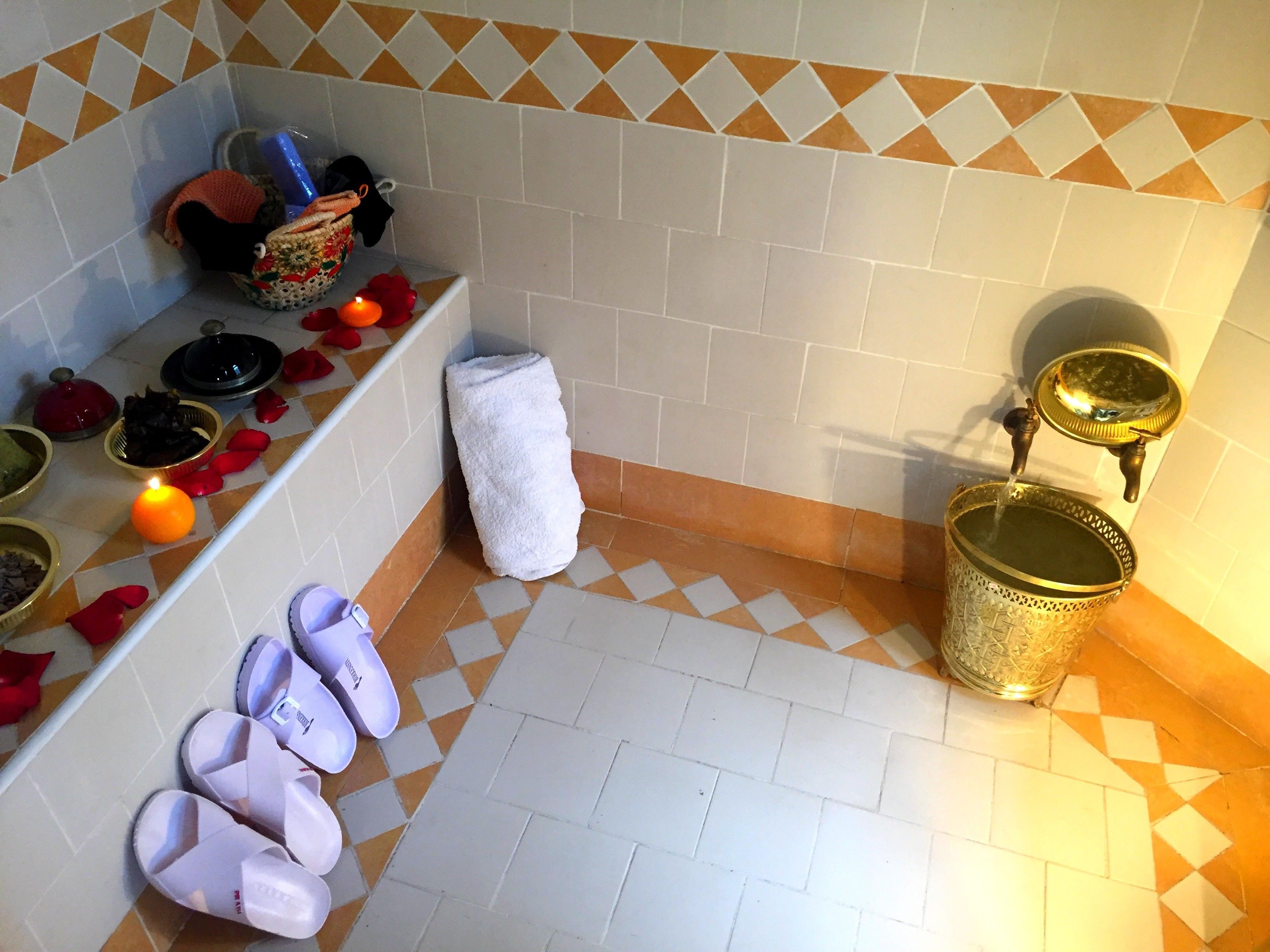 A bathroom with a tiled floor, a white towel, slippers, a basin, and a shelf with toiletries.