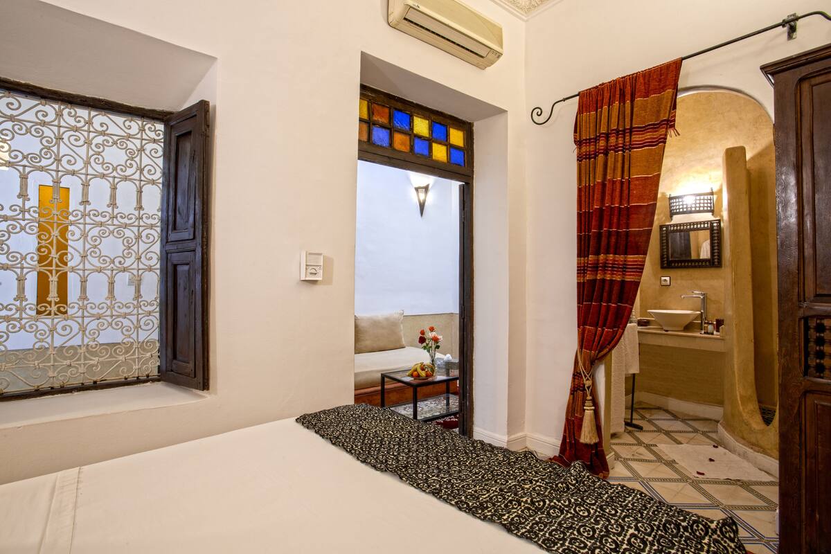 A hotel room with a bed, a window with decorative ironwork, a bathroom with a sink and mirror, and a patterned rug.