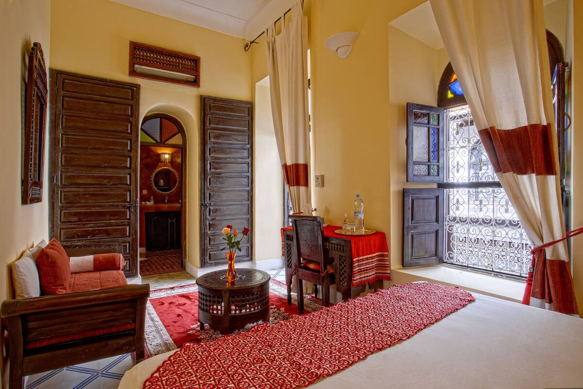 A room with a large bed, a wooden chair, a small table with a vase, and a window with curtains.