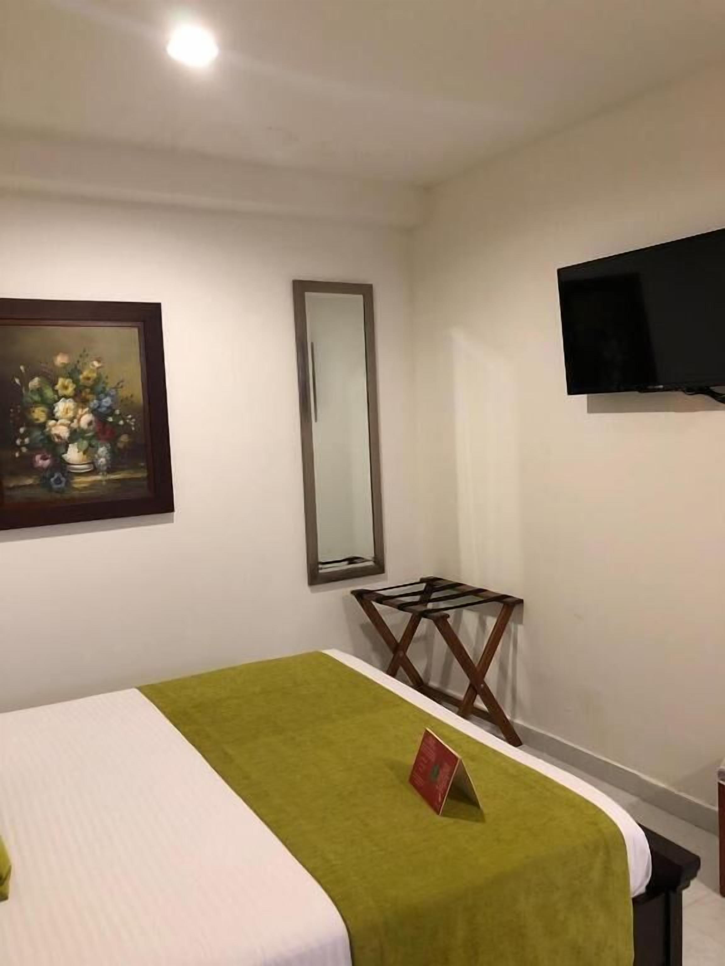 A hotel room with a bed, a painting, a mirror, a flat-screen TV, and a small table.