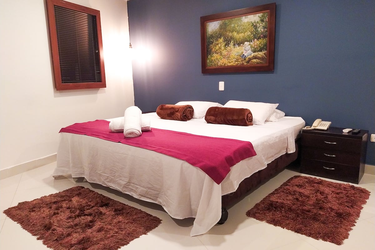 Una cama bien hecha con ropa de cama blanca y marrón, un cuadro en la pared y una mesita de noche con un teléfono y un mando a distancia.
