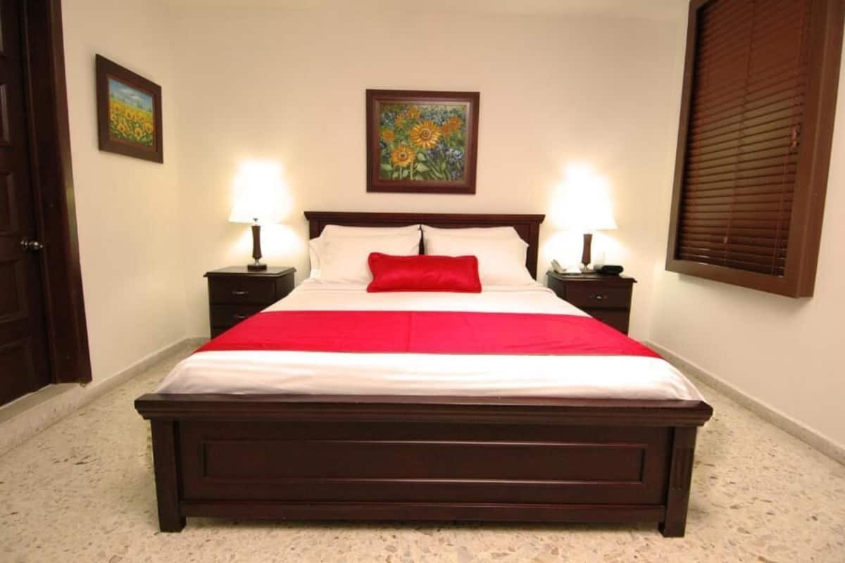 Una cama bien hecha con un cojín rojo decorativo, dos lámparas de noche y un cuadro con motivos florales encima.