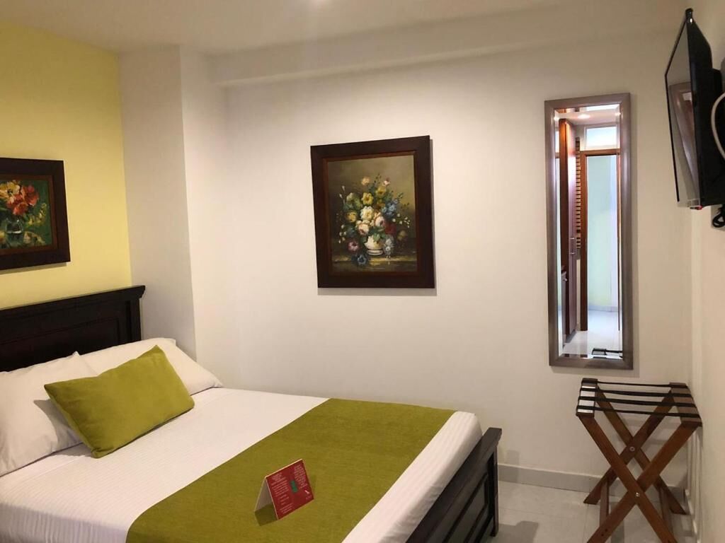 A hotel room with a bed, a green throw, a painting, a window, and a TV mounted on the wall.