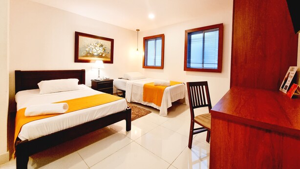 A hotel room with a large bed, a smaller bed, a desk, and a chair. There are two windows with blinds, a painting on the wall, and a lamp on the nightstand. - Hotel Karlo (Cali)