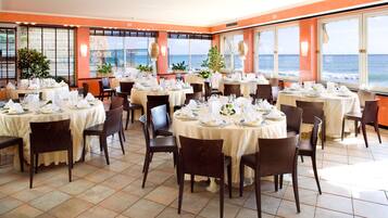 Un restaurant avec des tables rondes disposées pour le service, de grandes fenêtres offrant une vue sur l’océan et des plantes en pot.