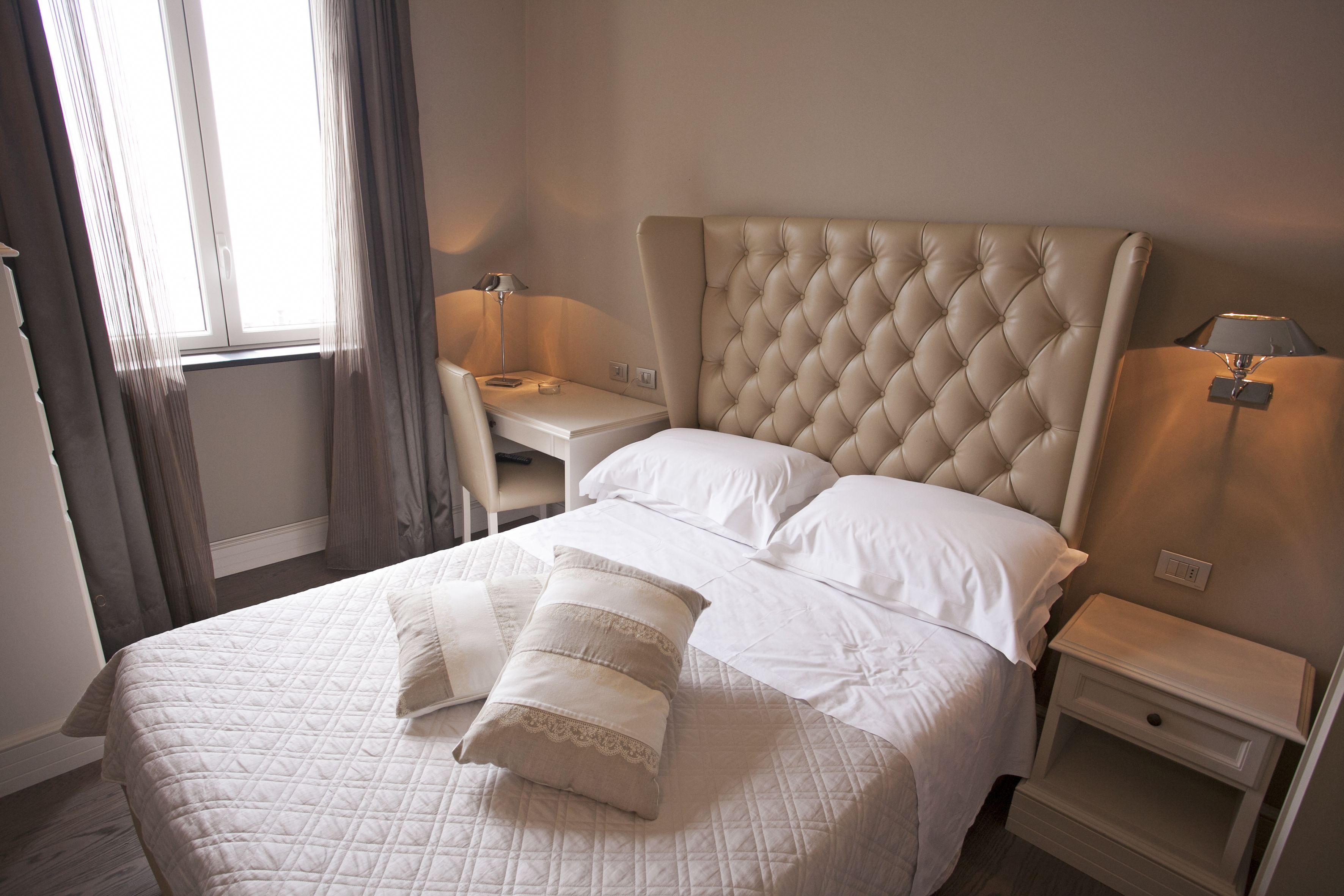A neatly made bed with a quilted cover, two bedside tables with lamps, and a window with curtains.