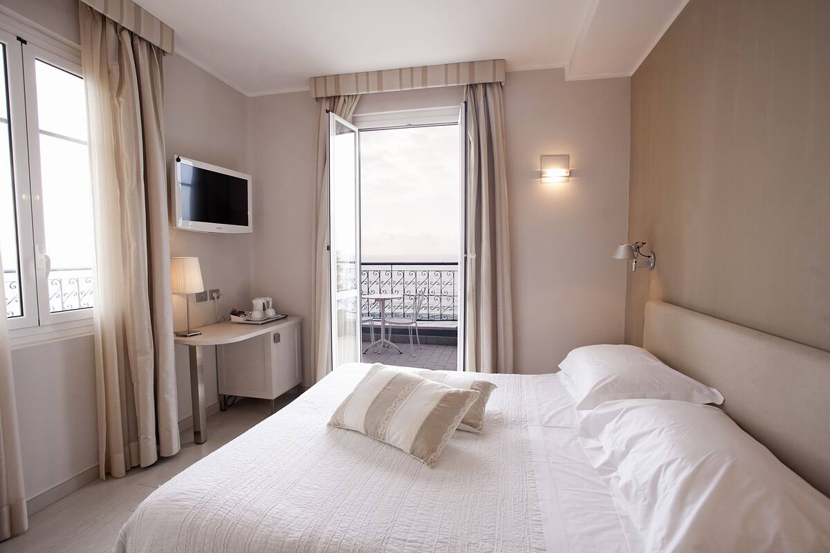A hotel room with a bed, a desk with a lamp, a TV, and a balcony with a table and chairs.
