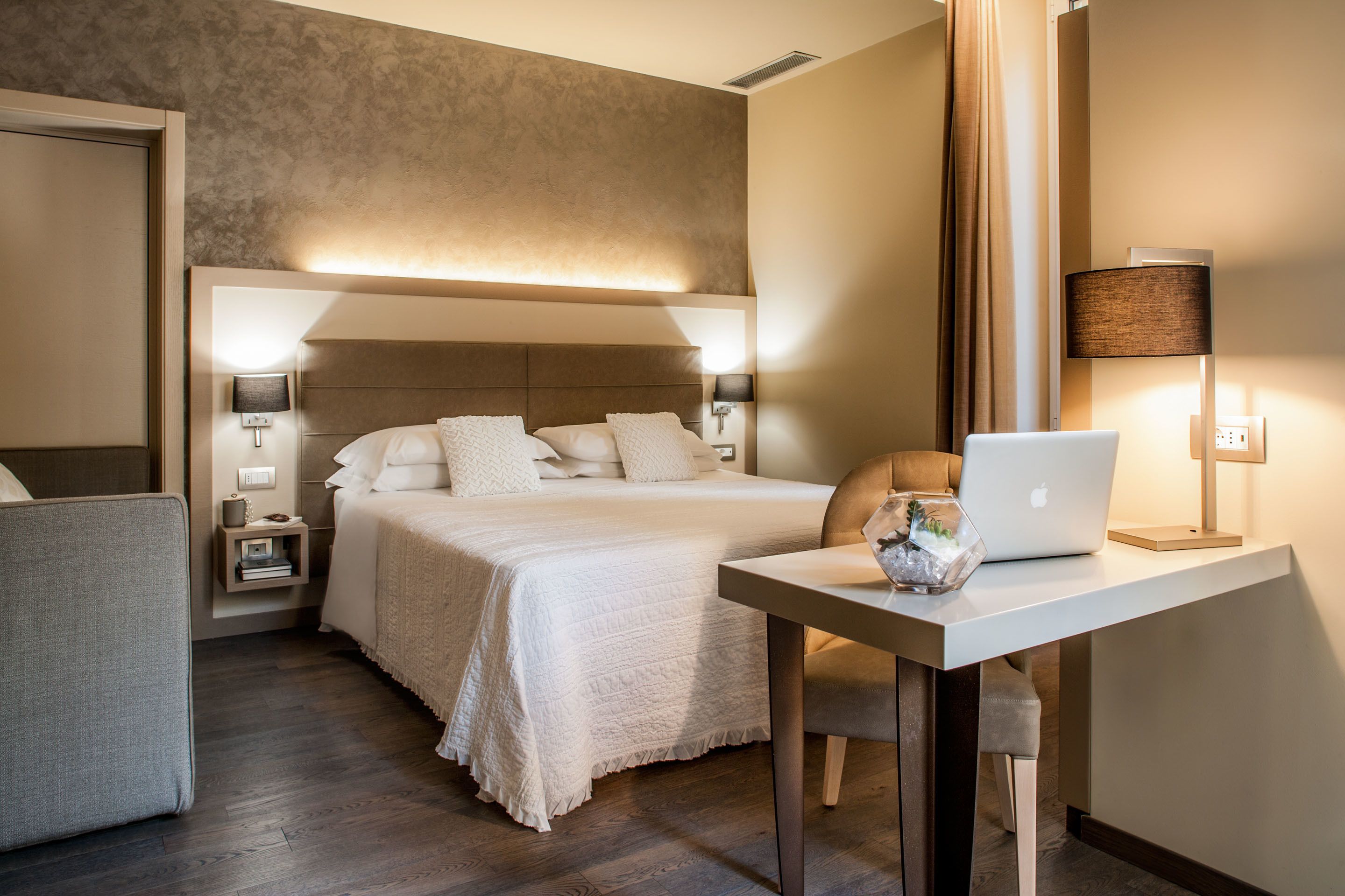 A modern hotel room with a large bed, a desk with a laptop, a chair, and a nightstand with a lamp.