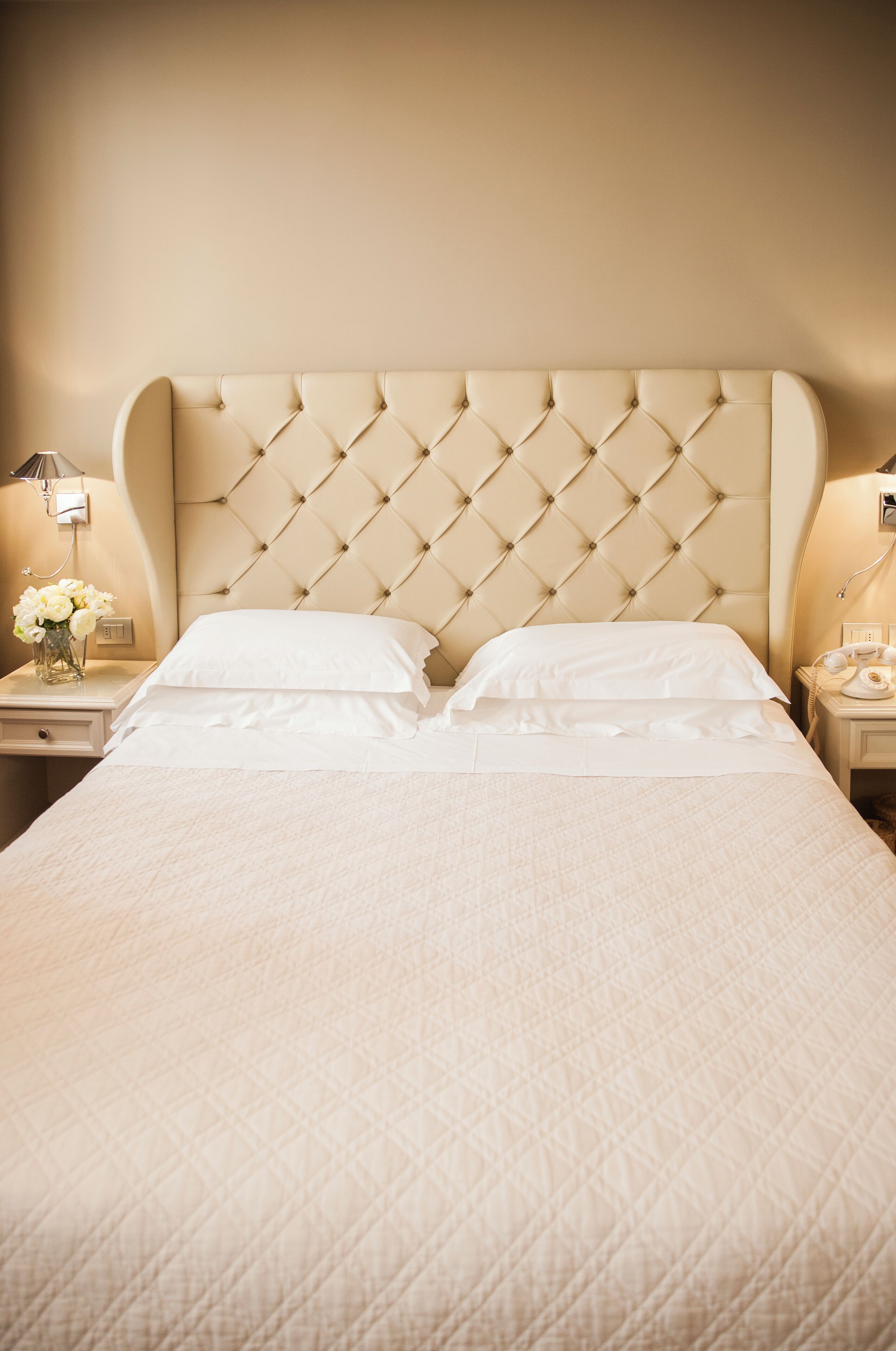 A neatly made bed with a tufted headboard, white linens, and a bedside table with a lamp and a vase of flowers.