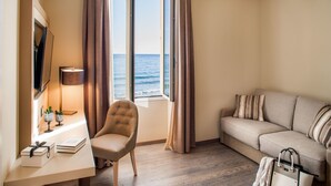 Habitación de hotel con suelo de madera, un sofá, una silla, un escritorio con lámpara, un televisor y una ventana con cortinas.
