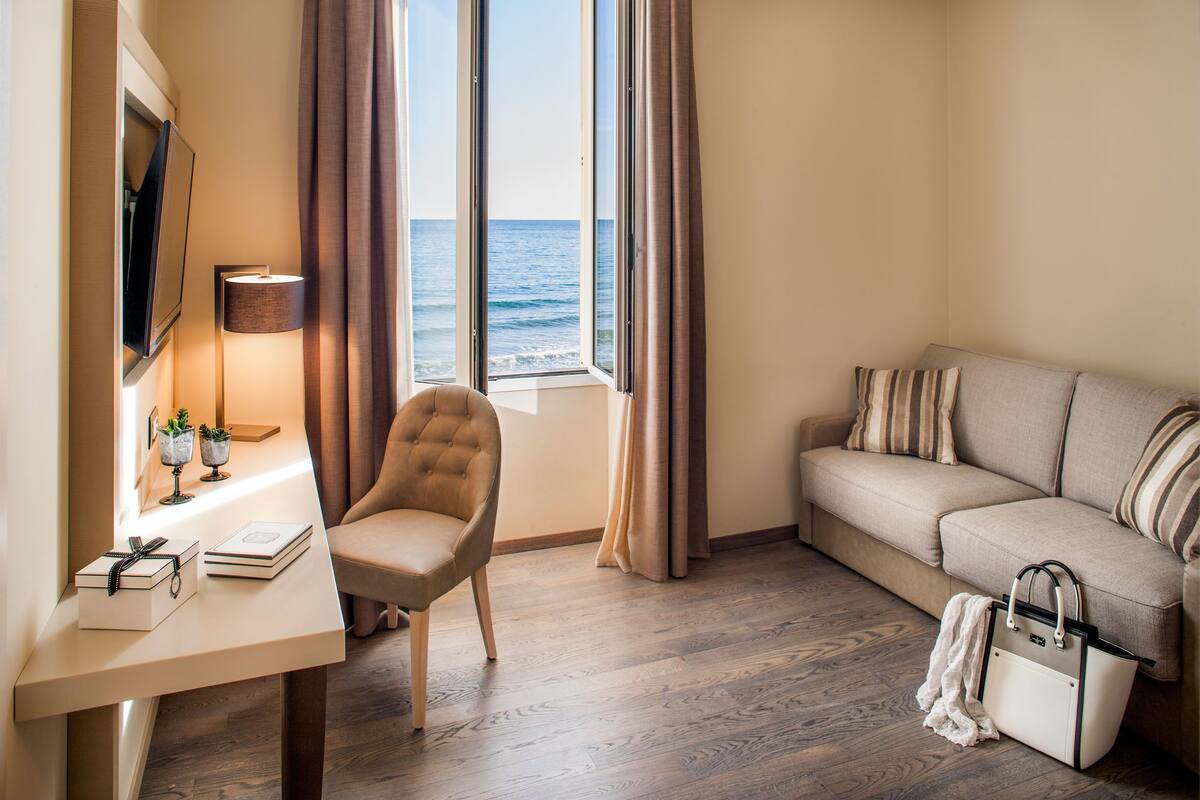 A hotel room with a wooden floor, a sofa, a chair, a desk with a lamp, a TV, and a window with curtains.